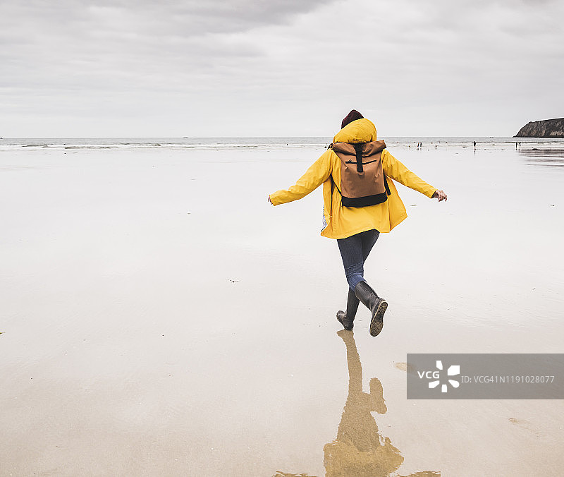 在法国布列塔尼海滩跑步的黄色雨衣年轻女人图片素材