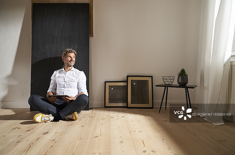 放松的中年男人坐在地板上，拿着平板电脑看着远方图片素材