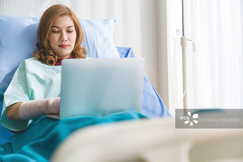 亚洲女人在病床上使用笔记本电脑和智能手机放松身心，住院康复图片素材