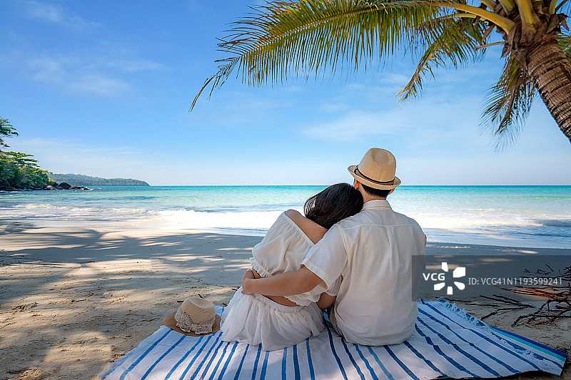 年轻的亚洲夫妇在热带海滩的棕榈树下相爱，眺望美丽的夏日海景，享受蜜月。图片素材