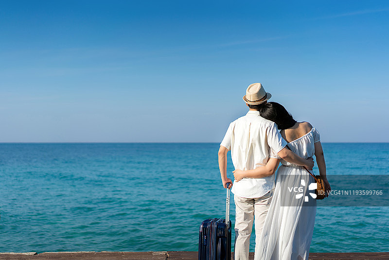 亚洲情侣在海滩木甲板上手持行李箱和背包欣赏海景的背影图片素材