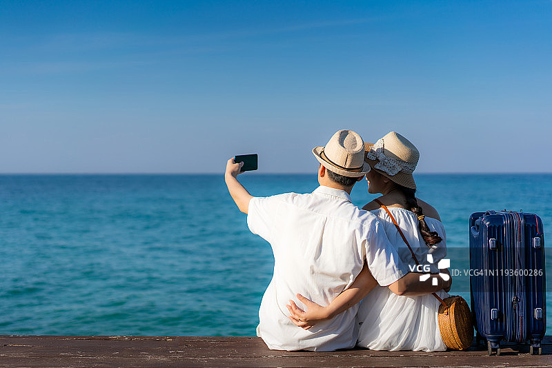 快乐的年轻亚洲情侣在泰国桐艾的木甲板海上用智能手机自拍。蜜月情侣在海边浪漫夏季旅行图片素材