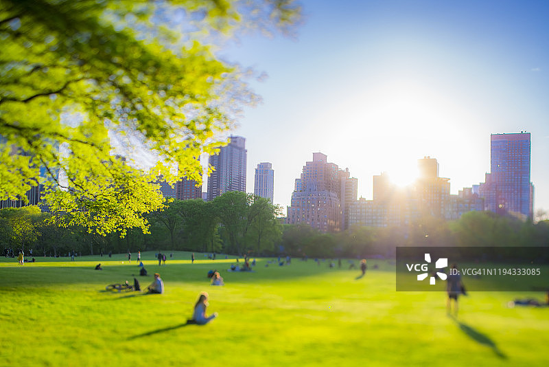 2019年5月11日，纽约中央公园西区沐浴在春日落日余晖中，照亮着建筑、绿树和绵羊草地上的人们图片素材