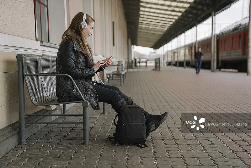 微笑的年轻女子背着背包坐在站台上使用智能手机和耳机，维尔纽斯，立陶宛图片素材