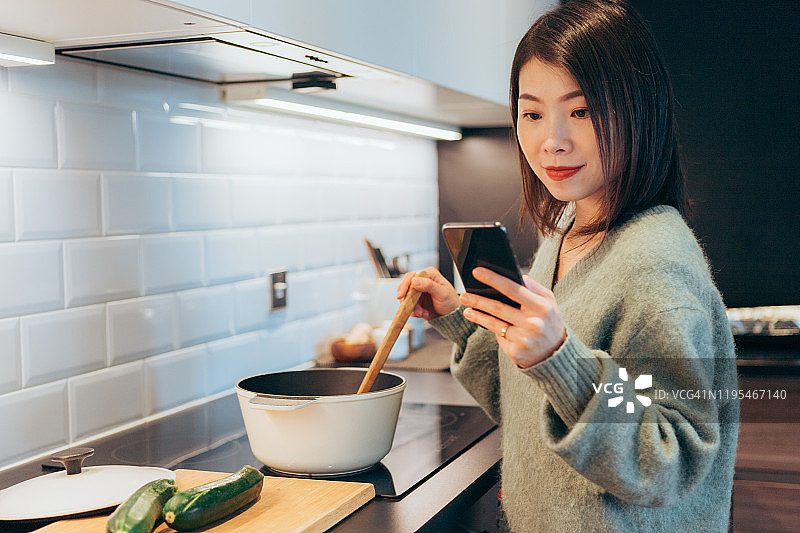 年轻快乐的美丽女人在家中一边做饭一边看手机上的食谱图片素材