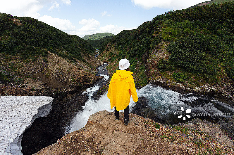 瀑布雪山背景下，身穿白色巴拿马帽和黄色雨衣的年轻女孩：夏季图片素材