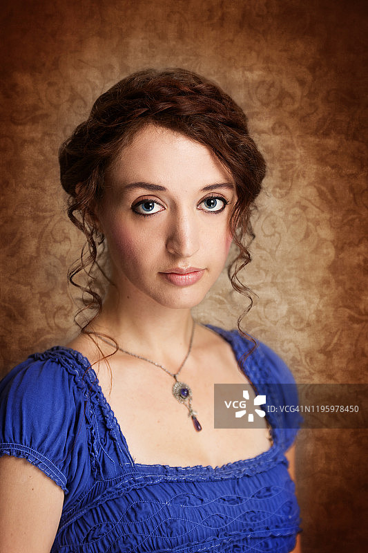 年轻红发女子盘着发髻面无表情地看着镜头的影棚肖像，佩戴吊坠项链，身穿方形领口上衣，背景为金棕色织锦图片素材