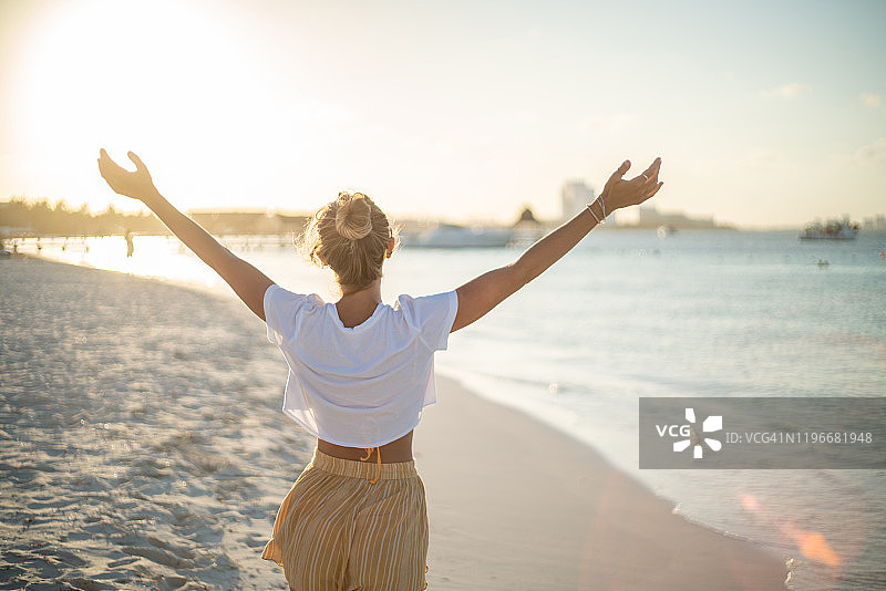 快乐的年轻女人在日落时拥抱大自然；女性站在海滩上伸出双臂图片素材