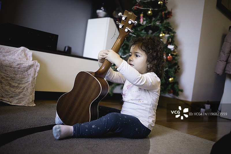 小女孩在圣诞树背景下检查她的新尤克里里琴图片素材