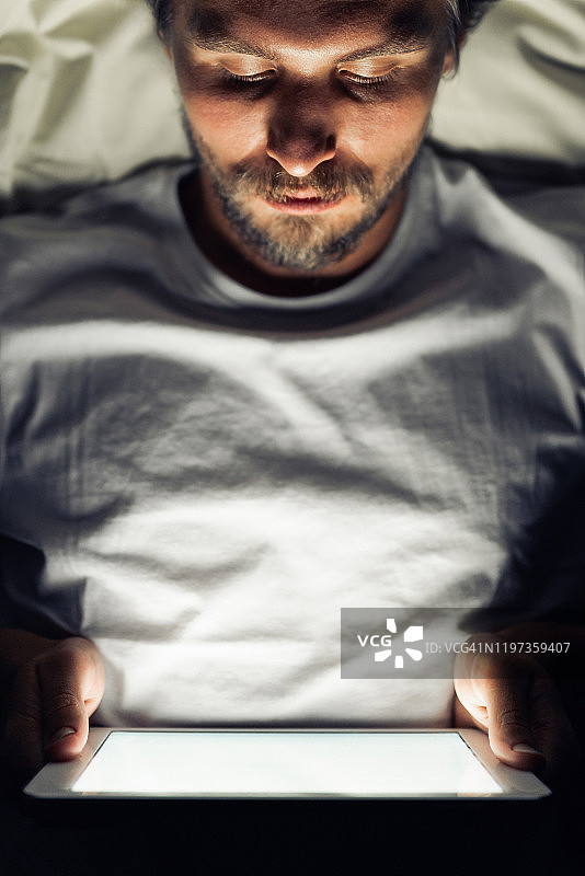 年轻英俊、疲惫的男子，留着胡子，晚上看着平板电脑图片素材