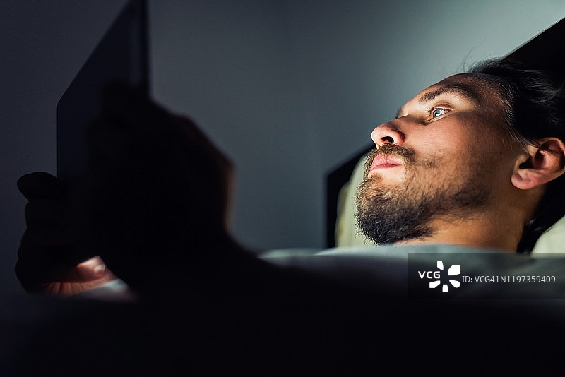年轻英俊、疲惫的男子，留着胡子，晚上看着平板电脑图片素材