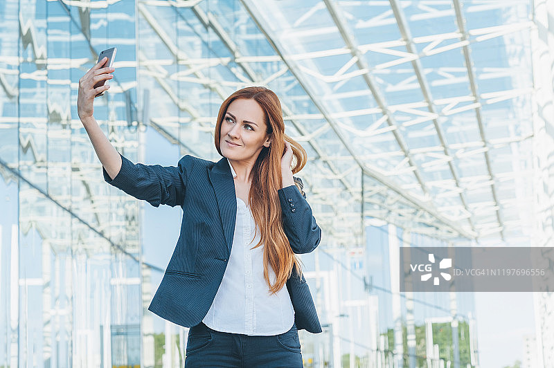 在现代玻璃商务中心背景下，手持电话的商务女孩自拍图片素材