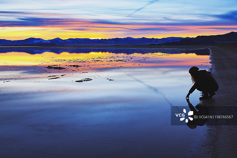 年轻女子蹲在湖边触摸水面，日落时分地平线上有山脉的轮廓和倒影，犹他州博纳维尔盐滩图片素材