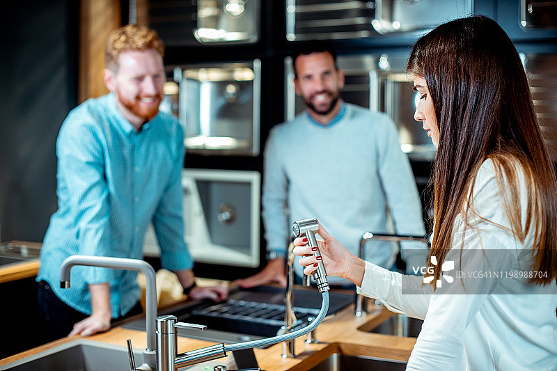 年轻夫妇在厨房设备商店里，在店员的帮助下，检查带冲洗软管的洗碗池水龙头图片素材