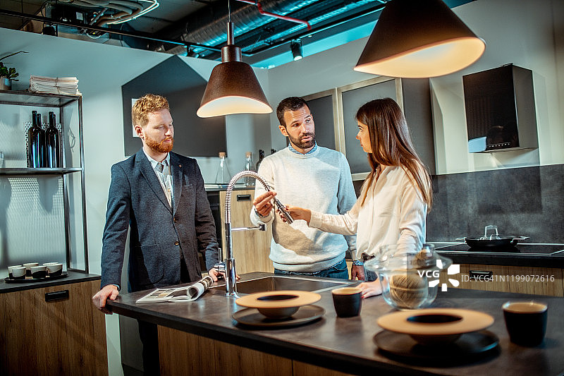 年轻夫妇在厨房设备商店检查洗碗池水龙头的冲洗软管功能，商店销售员在一旁协助图片素材