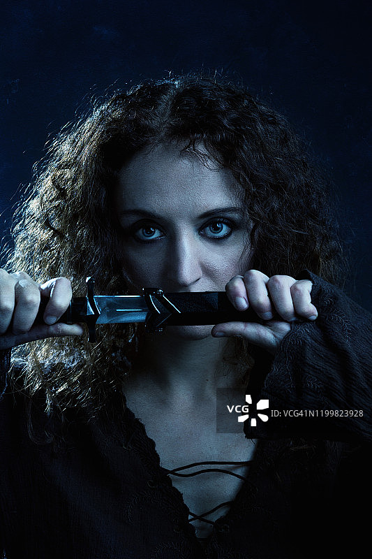 有着深色卷发的年轻女人拔出匕首直视镜头，冷色调工作室拍摄图片素材