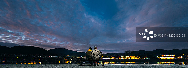 在黄昏时分，参观日本京都东山传统小镇，坐在长椅上欣赏湖边日落的年长夫妇背影图片素材