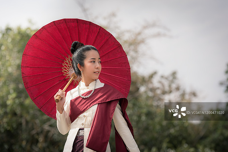 穿着传统东北服装、手持红伞的伊善美女，泰国女士穿着当地服装的肖像，东南亚文化图片素材
