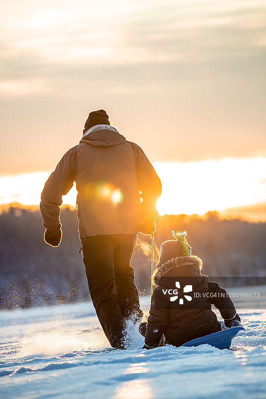 日落时分，爷爷在冰冻的湖面上拉着孙子在雪橇上玩耍图片素材
