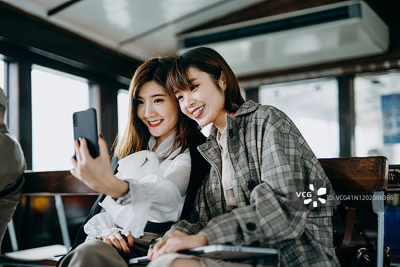 两个微笑的年轻亚洲女人坐在渡轮上用智能手机自拍图片素材