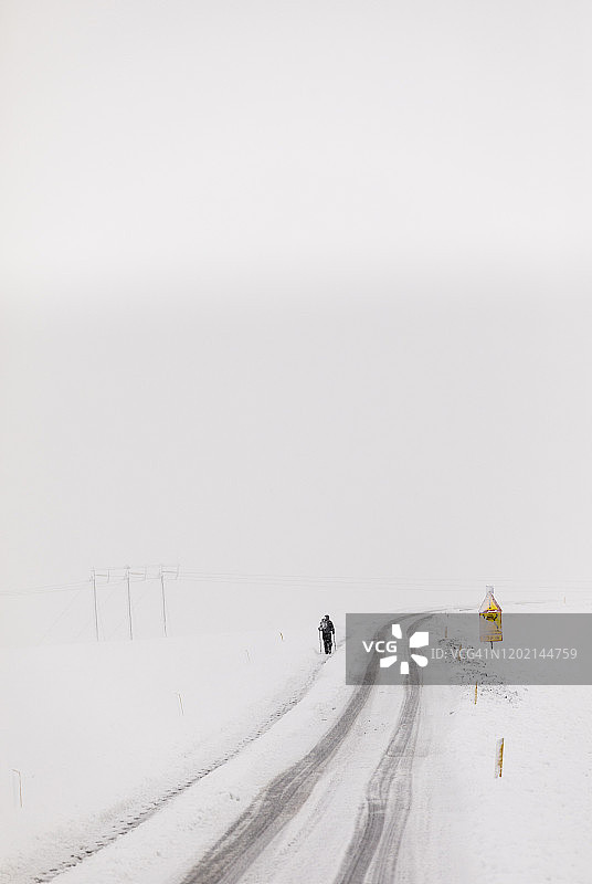 在冰岛高速公路边缘，手持滑雪杖行走的男人，空旷的高速公路被风雪覆盖图片素材