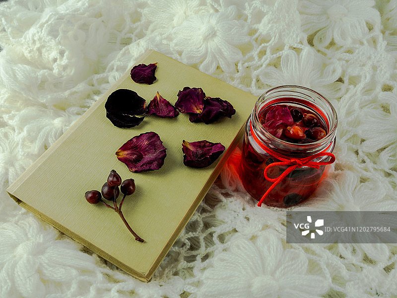 玻璃杯中红色水果、玫瑰果、沙棘和玫瑰花瓣的红色混合物，旁边是带有红色蝴蝶结的书，放在毯子上图片素材