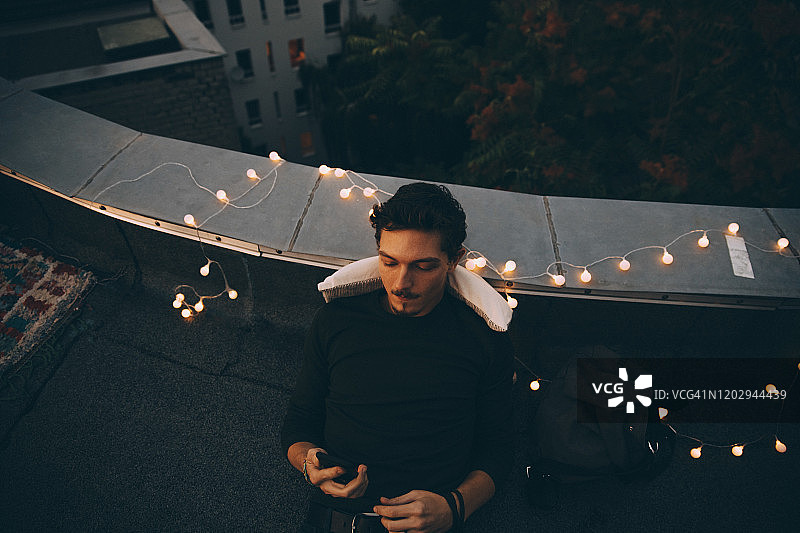 黄昏时分在灯光照亮的露台上放松身心并使用智能手机的年轻男子高角度视图图片素材