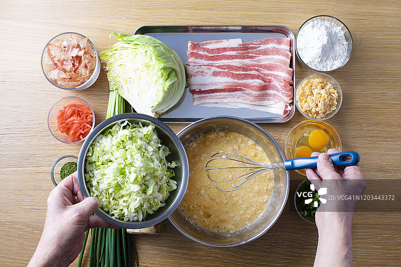 日本御好烧的配方。山药、面粉、白菜、猪肉、天须、鸡蛋、红姜、木鱼、蓝海藻、青葱等食材摆放在木纹桌上。我奶奶把卷心菜切成美味的蛋糕、鸡蛋、面粉、山药和污渍图片素材