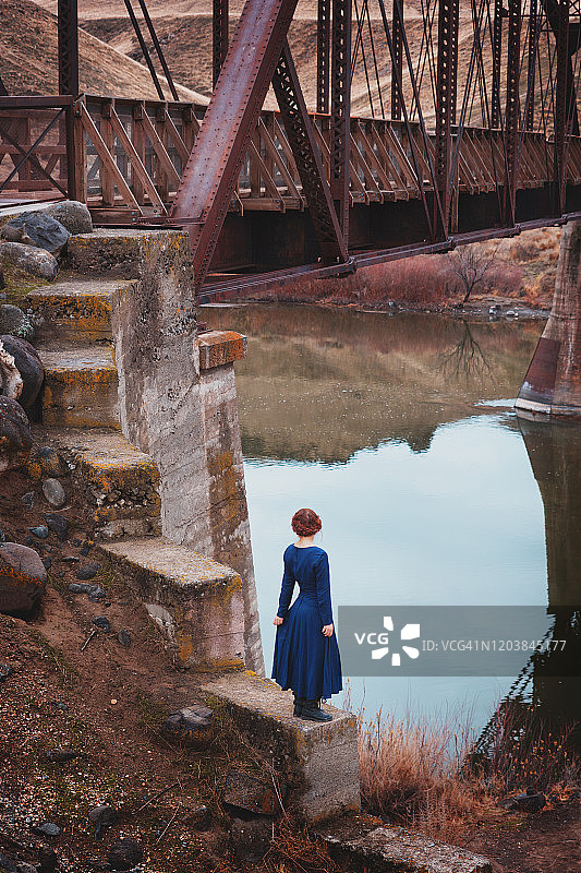 在老铁路桥底的墙上，一位扎着红色辫子，穿着蓝色复古长袖连衣裙的年轻女子背影，俯瞰河流图片素材