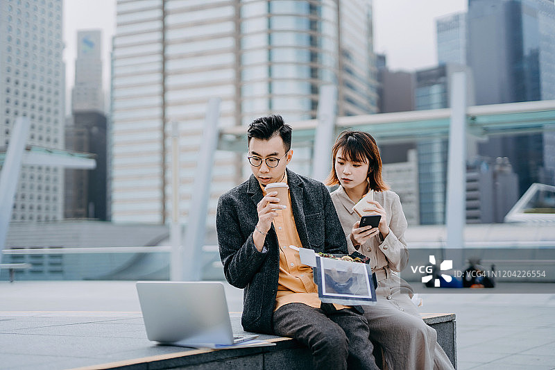 亚洲商业同事在城市阳台上一起午餐，使用笔记本电脑和智能手机工作。图片素材