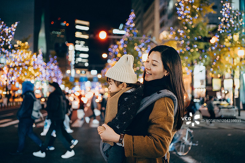 年轻母亲和小女儿在夜间城市街道上行走，背景是灯火通明的城市建筑图片素材