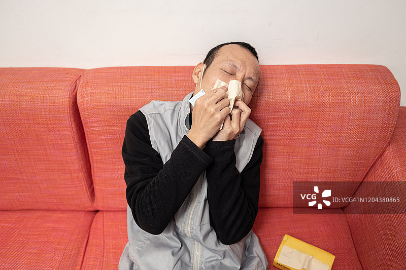 亚洲男人坐在沙发上用冷毛巾擦鼻子，他很虚弱图片素材