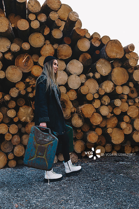 长发金发女子，戴黑色羊毛帽，穿黑色羊毛外套、黑色长袜和白色靴子，手提汽油桶，站在木柴堆后图片素材