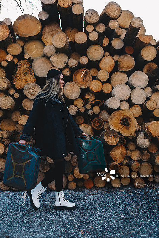 长发金发女子，戴黑色羊毛帽，穿黑色羊毛外套、黑色长袜和白色靴子，手提汽油桶，站在木柴堆后图片素材