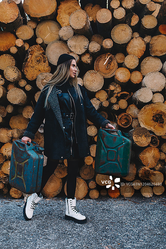 长发金发女子，戴黑色羊毛帽，穿黑色羊毛外套、黑色长袜和白色靴子，手提汽油桶，站在木柴堆后图片素材