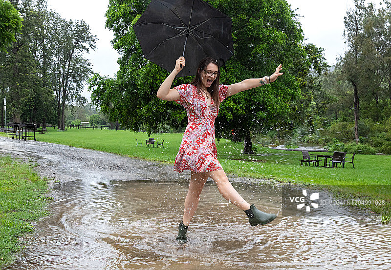 穿着红色连衣裙和靴子，拿着黑色雨伞在水坑里跳舞的年轻女人图片素材