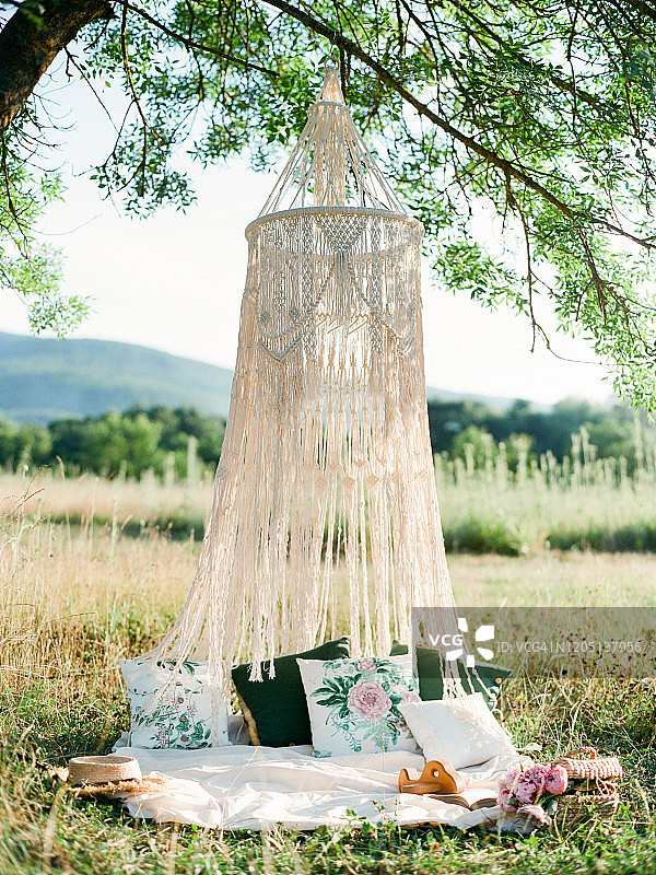 挂在大树上的大型棉质流苏华盖，树荫下带有枕头和鲜花的波西米亚野餐，浪漫周末创意图片素材