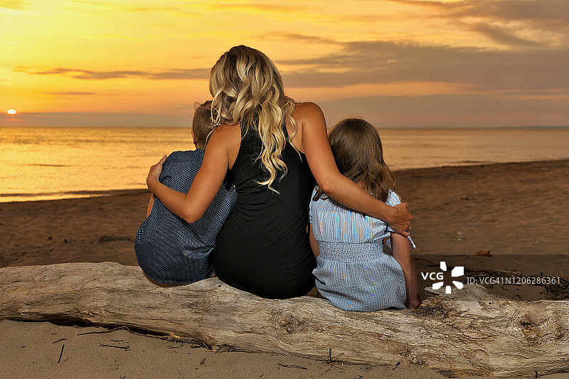 一位年轻母亲和她的青少年儿子和女儿一起坐在日落时海滩的浮木上，后视图图片素材