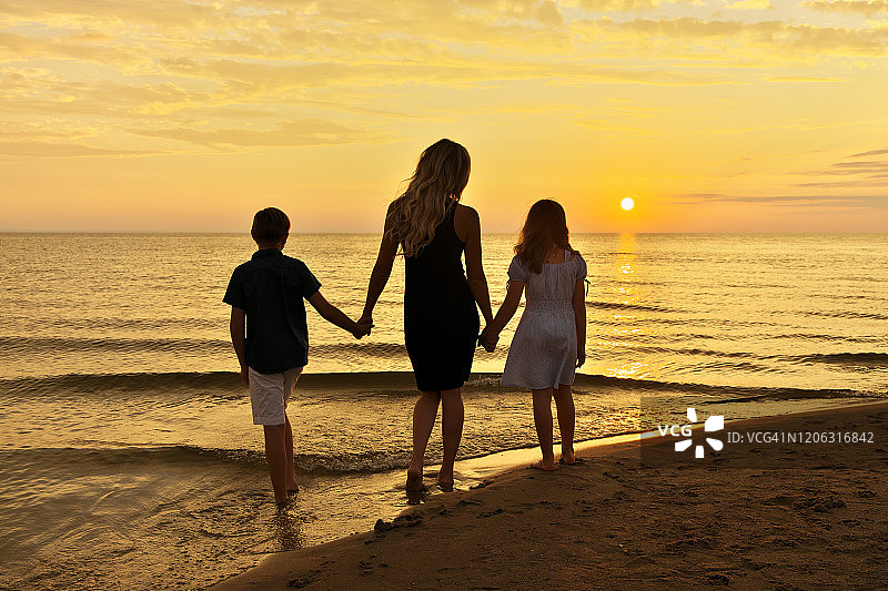 年轻的母亲和她十几岁的儿子和女儿手牵着手在海滩观看壮丽日落的剪影，后视图图片素材