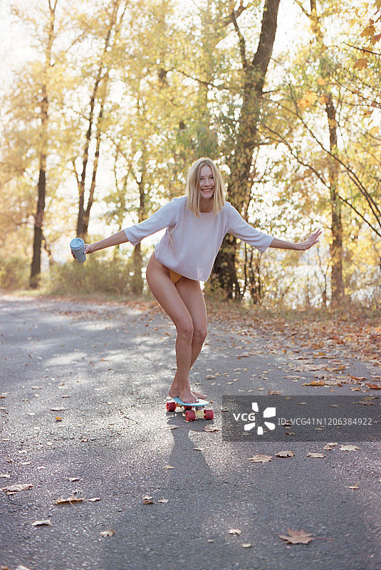 年轻金发女子拿着一个环保咖啡杯，在秋天的森林里滑着滑板。图片素材