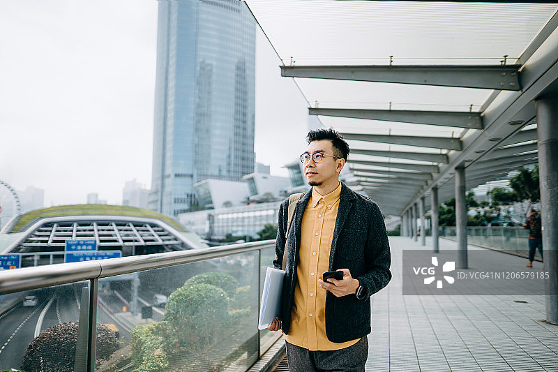 在城市通勤的亚洲商务男士肖像，肩背背包使用智能手机图片素材