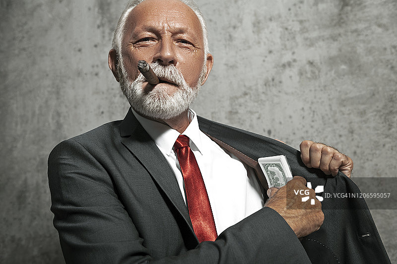 穿着经典西装，抽着雪茄的成熟胡须商人正把钱放进外套内袋图片素材