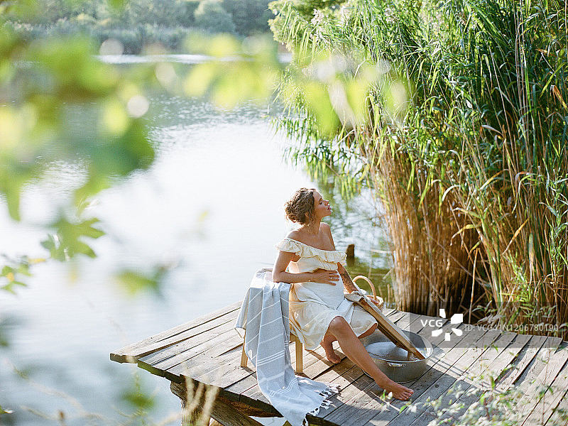 穿着复古连衣裙的年轻孕妇在河边金属盆里清洗亚麻布，孕妇复古海报风照片，生态洗衣概念图片素材
