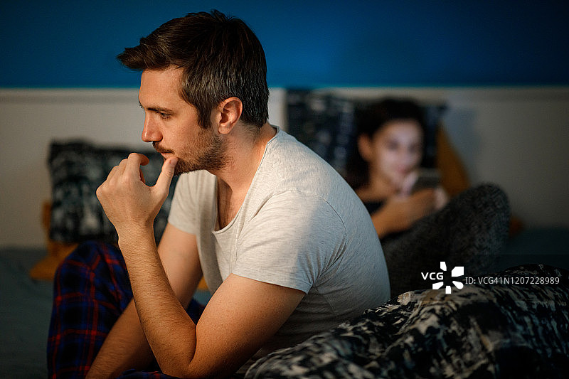 晚上，焦虑的男人坐在床边，妻子在使用手机图片素材