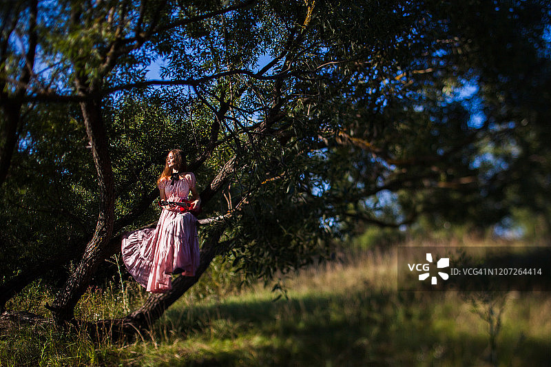 穿着粉色连衣裙、戴着帽子的漂亮女孩站在绿树旁图片素材