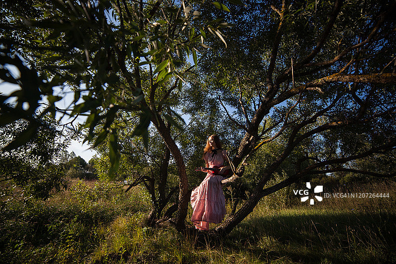 穿着粉色连衣裙、戴着帽子的漂亮女孩站在绿树旁图片素材