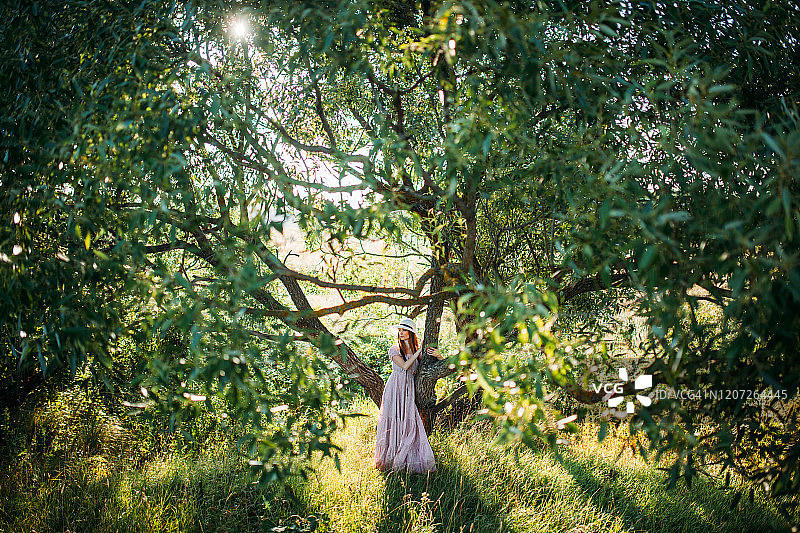 穿着粉色连衣裙、戴着帽子的漂亮女孩站在绿树旁图片素材