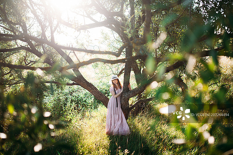 穿着粉色连衣裙、戴着帽子的漂亮女孩站在绿树旁图片素材