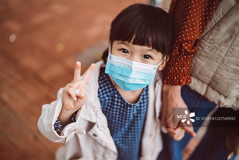 戴着医用口罩在街上对着镜头欢快地比V字手势的小女孩图片素材