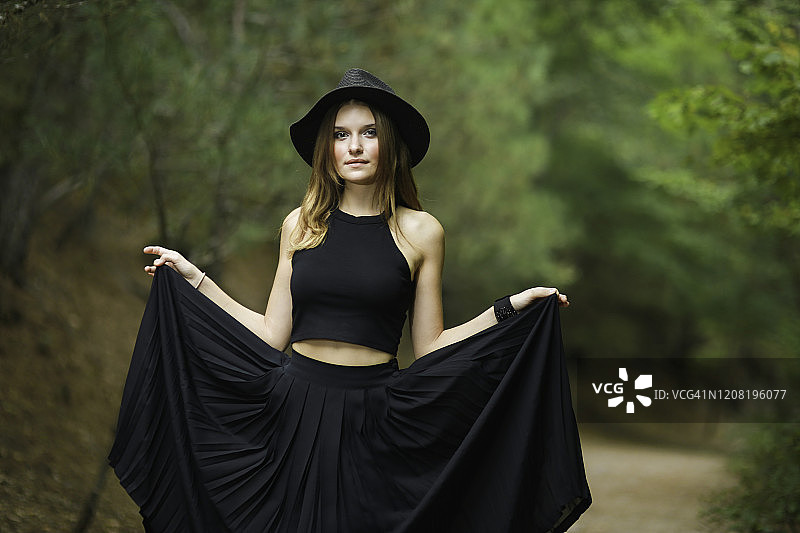 戴着帽子、穿着黑色连衣裙的金发年轻女子在树林中的肖像。有魅力的女孩在散步。现代女性时尚图片素材
